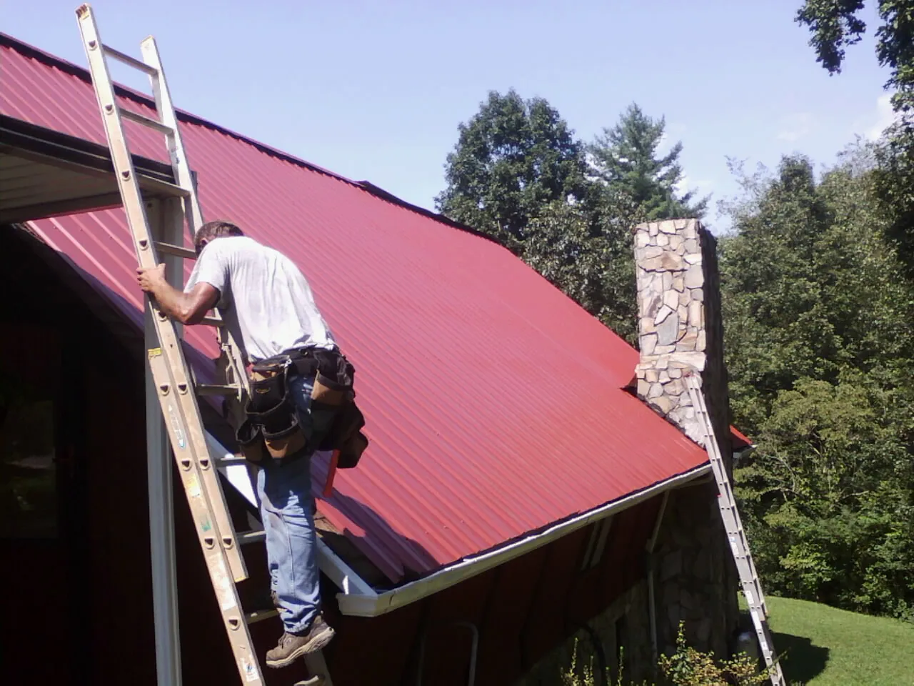 Roofer on a ladder beside a red metal roof and stone chimney — Soffit & Fascia Repair by Roofing Brothers in Columbia Falls